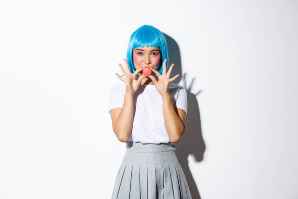 Coquettish asian girl in blue wig showing macaron near lips and smiling, standing over white background - Photo, Image