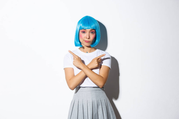 Нерешительная милая азиатка в голубом парике, указывающая пальцами в сторону с озадаченным выражением лица, делающая выбор, стоящая на белом фоне - Фото, изображение