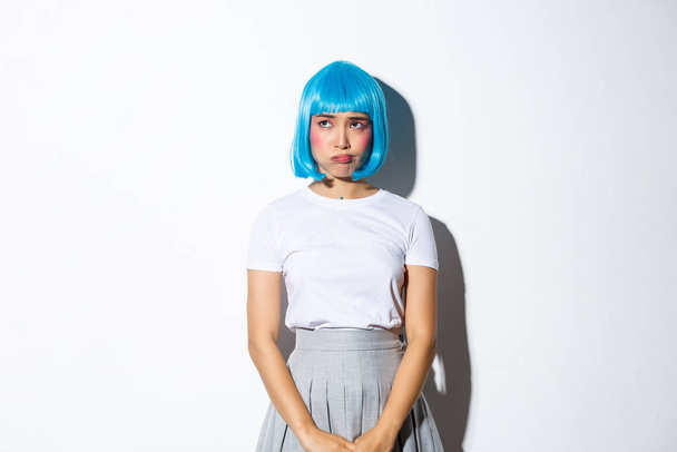 Porträt eines komplizierten, traurigen asiatischen Mädchens mit blauer Perücke, schmollend und mit beunruhigtem Gesichtsausdruck in die linke obere Ecke schauend, vor weißem Hintergrund stehend - Foto, Bild