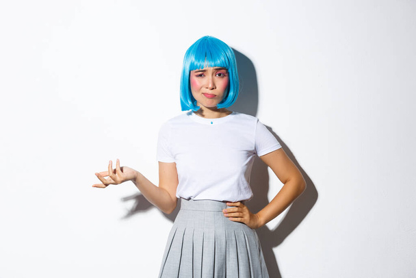 Изображение скептически настроенной азиатской девушки в голубом парике, выглядящей смущенной и осуждающей, стоящей на белом фоне - Фото, изображение