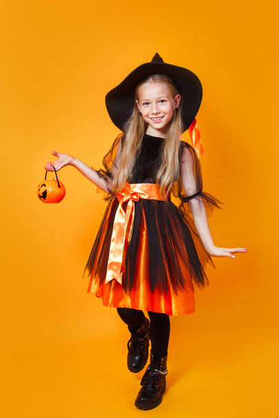 Cute happy little girl dressed up as witch with bucket of candy on orange background - Fotó, kép
