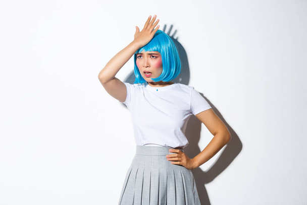 Portrait of bothered asian girl in blue party wig, slap forehead and looking pissed-off, forgot something, standing in halloween costume over white background - Foto, Bild