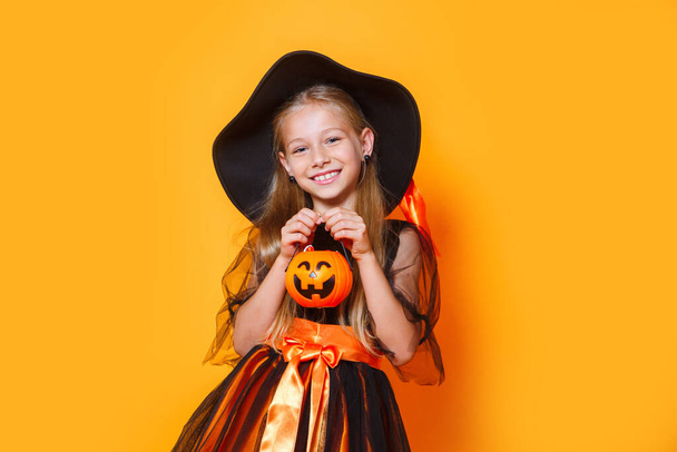 Маленькая девочка, одетая как ведьма и держащая корзину с тыквой на оранжевом фоне - Фото, изображение