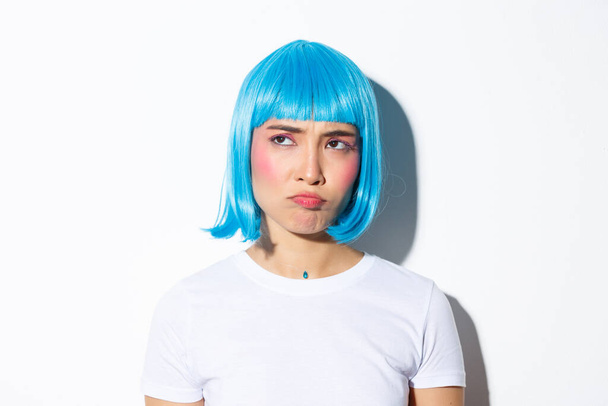 Imagen de linda chica asiática en peluca azul mirando decepcionada o celosa, enfurruñada mientras mira fijamente a la esquina superior izquierda, de pie sobre fondo blanco - Foto, Imagen
