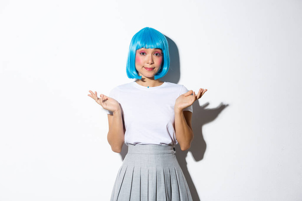 Porträt eines ahnungslosen jungen asiatischen Partygirls mit blauer Perücke, das achselzuckend, unentschlossen in die Kamera blickt und vor weißem Hintergrund steht - Foto, Bild