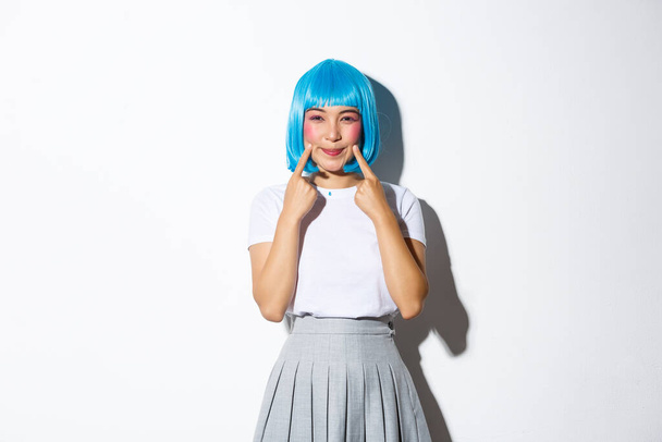 Mavi peruklu, gülümseyen ve gamzelerine işaret eden, beyaz arka planda duran Cadılar Bayramı partisi için giyinmiş Kawaii Asyalı bir kız resmi. - Fotoğraf, Görsel
