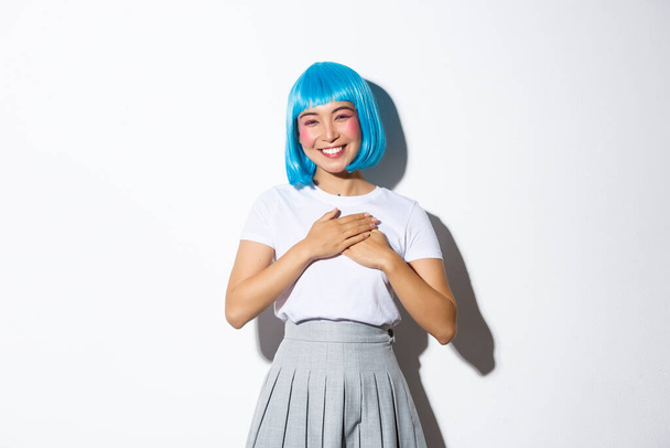 Image of thankful smiling asian girl in halloween costume and blue party wig, holding hands on heart and looking grateful, thanking for something, standing over white background - Photo, Image