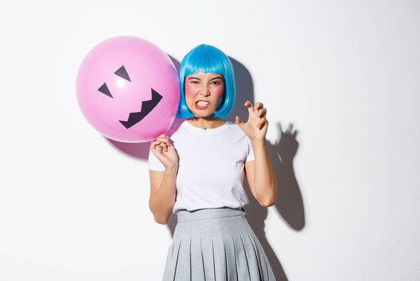 Portret van een grappig Aziatisch meisje dat iemand bang probeert te maken op Halloween, een blauwe pruik draagt en een roze ballon vasthoudt met een eng gezicht - Foto, afbeelding