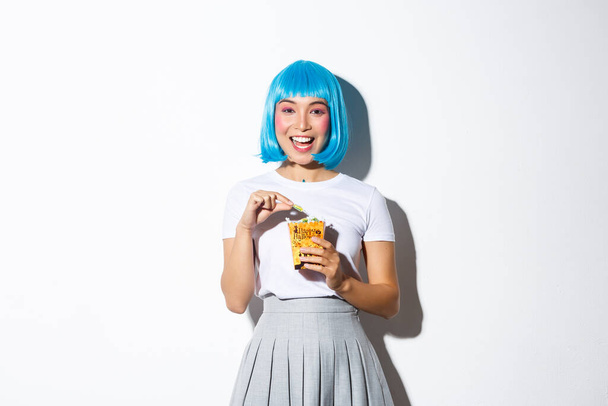 Afbeelding van schattig aziatisch meisje vieren halloween, het eten van snoep uit truc of traktatie zak, het dragen van blauwe korte pruik en anime schoolmeisje kostuum, staande over witte achtergrond - Foto, afbeelding