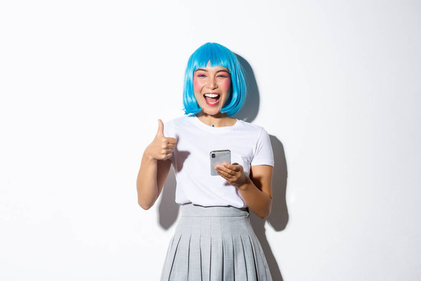 Portrait de jolie femme asiatique en perruque bleue riant heureux, montrant pouces levés, tenant téléphone portable - Photo, image