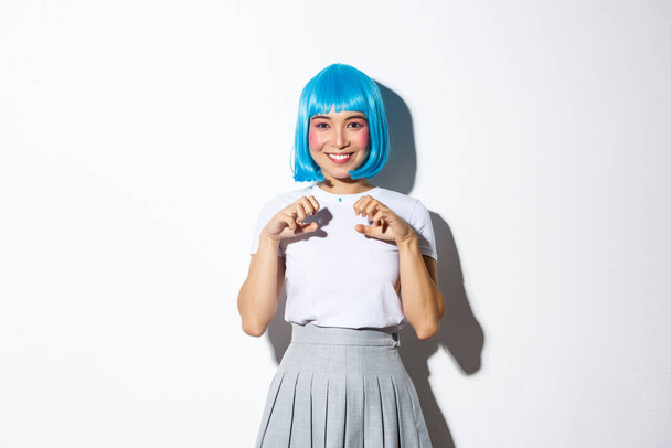 Πορτρέτο του όμορφου κοριτσιού από την Ασία με μπλε περούκα, απολαμβάνοντας αποκριές, χαμογελώντας και διασκεδάζοντας στο πάρτι, στέκεται πάνω από το λευκό φόντο - Φωτογραφία, εικόνα