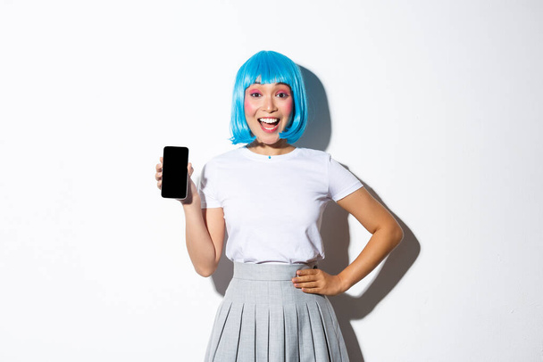 Retrato de chica asiática bonita feliz en peluca azul y disfraz de halloween, mostrando la pantalla del teléfono inteligente y sonriendo alegremente, de pie sobre fondo blanco - Foto, Imagen