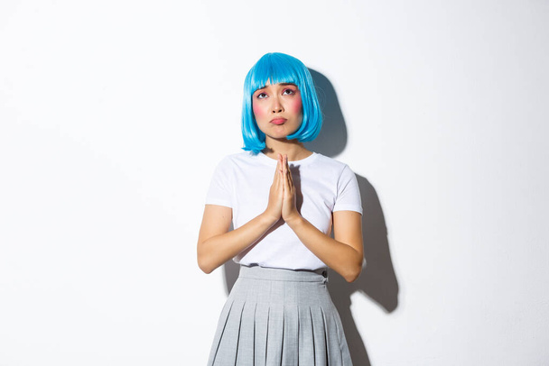 Portrait of hopeful sad girl looking up and praying or begging for God help, standing in blue wig over white background - Foto, afbeelding