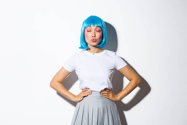 Портрет милой милой азиатской девушки, закрытые глаза и дуться для поцелуя, носить синий парик и костюм школьницы, стоя на белом фоне - Фото, изображение