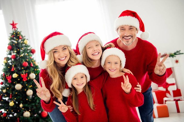 Photo portrait of funny friendly family wearing warm red festive jumpers and headwear preparing for christmas eve near xmas tree decorations showing v-sign with two fingers - Foto, imagen