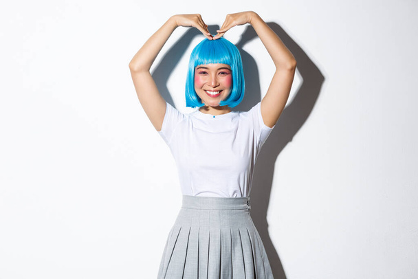 retrato de bonito japonês menina no azul peruca mostrando coração gesto e sorrindo, de pé sobre branco fundo - Foto, Imagem