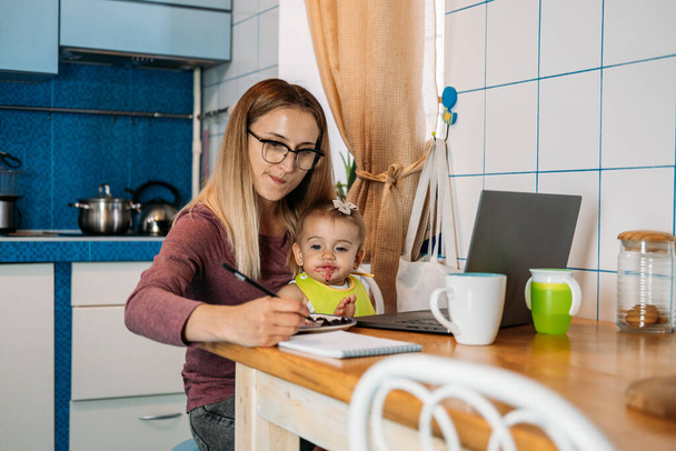 Evden çalışıyorum. Evde çalışan genç bir anne, mutfak arka planında dizüstü bilgisayar kullanıyor. Bebeğini besleyen, cep telefonuyla konuşan, iş yerinde dizüstü bilgisayarına bakan genç bir kadın.. - Fotoğraf, Görsel