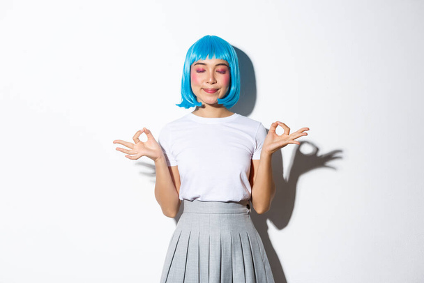 Portrait of beautiful asian girl in blue wig meditating, looking peaceful and smiling with eyes closed, standing in halloween costume over white background - Фото, зображення