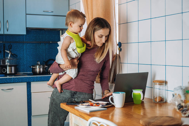 Arbeit von zu Hause aus. Junge Mutter mit Baby arbeitet zu Hause mit Laptop auf Küchenhintergrund Junge Frau füttert ihr Baby, telefoniert mit Handy, schaut auf Laptop am heimischen Arbeitsplatz. - Foto, Bild