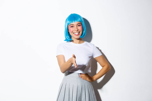 Friendly cute asian girl in blue wig greeting someone with pleasant smile, giving handshake and saying hello, standing over white background in halloween costume - Photo, Image
