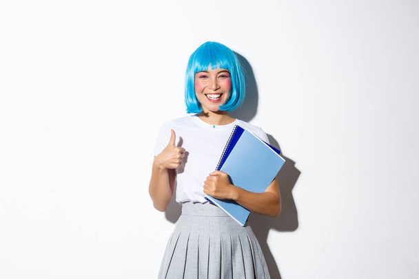 Mavi anime peruk takan, elinde okul defterleri olan, cadılar bayramı partisi için okul kızı gibi giyinmiş, onaylanmış, tatmin olmuş bir şekilde duran sevimli Asyalı kız resmi. - Fotoğraf, Görsel