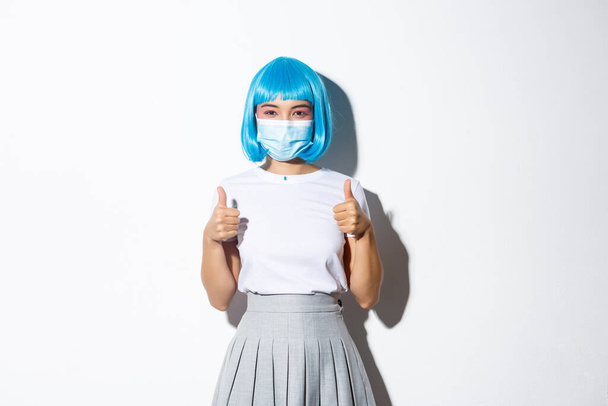 Concepto de celebración de Halloween y pandemia covid-19. Confiado joven asiática chica mostrando thumbs-up, usando médico máscara y azul peluca - Foto, imagen