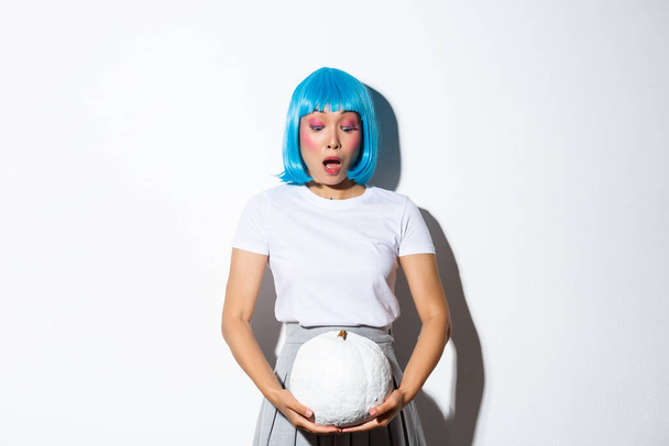 Image of surprised asian girl in blue wig, gasping and looking amazed at white pumpkin, celebrating halloween, standing over white background - Photo, Image
