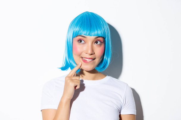 Bild von nachdenklichen niedlichen asiatischen Mädchen in blauer Perücke, Blick auf die obere linke Ecke und Denken, steht über weißem Hintergrund - Foto, Bild