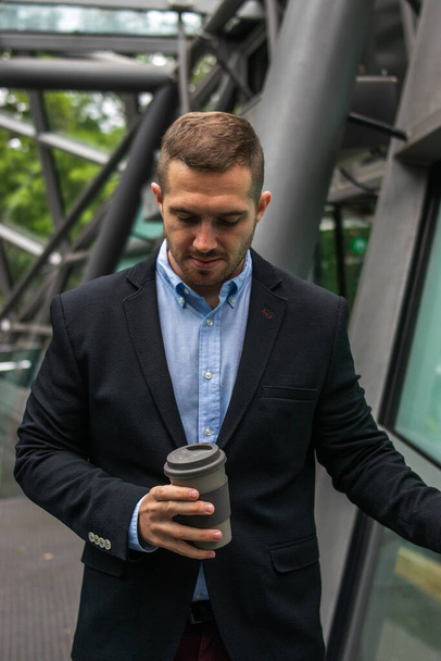 若い魅力的なビジネスマンが屋外でコーヒーブレイクを楽しんでいる写真。彼は賢い服を着ていて再利用可能なカップで飲んでいる - 写真・画像