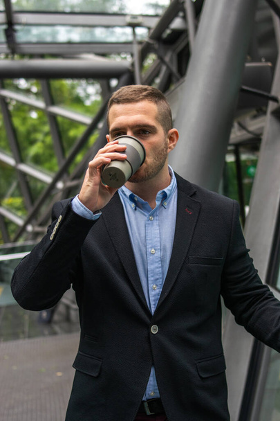 若い魅力的なビジネスマンが屋外でコーヒーブレイクを楽しんでいる写真。彼は賢い服を着ていて再利用可能なカップで飲んでいる - 写真・画像