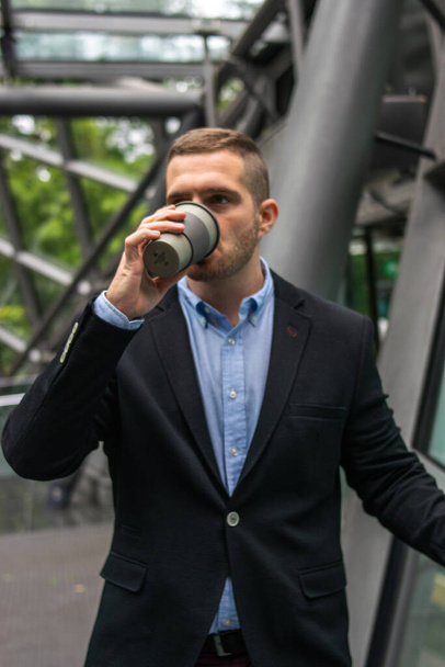 Фото молодого и привлекательного бизнесмена, наслаждающегося кофе-брейком на открытом воздухе. Он одет в умную одежду и пьет в чашке многоразового использования - Фото, изображение
