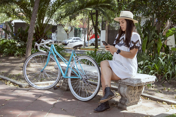 Η νεαρή Λατίνα τσεκάρει το κινητό της καθισμένη σε ένα πάρκο με ένα μπλε ποδήλατο δίπλα της. - Φωτογραφία, εικόνα