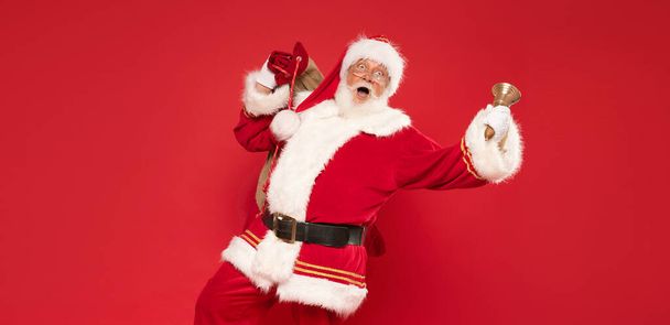 Happy, real, funny Santa Claus holding sack bag with Merry Christmas presents , delivering xmas gifts. Red background, a lot of copy space. - Photo, Image