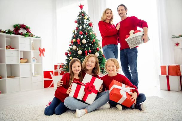 Photo of full big family five people gathering three small kids hold presents cuddle parents dad hold bag toys wear red jumper jeans in home living room many present boxes indoors - Фото, изображение