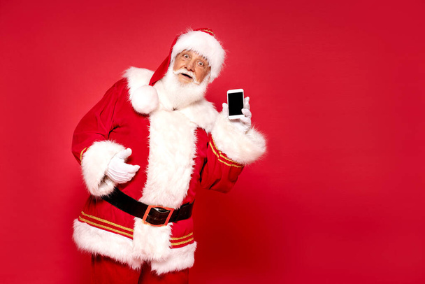 Babbo Natale davvero divertente che mostra lo schermo vuoto del telefono cellulare alla fotocamera. posa su sfondo rosso. - Foto, immagini