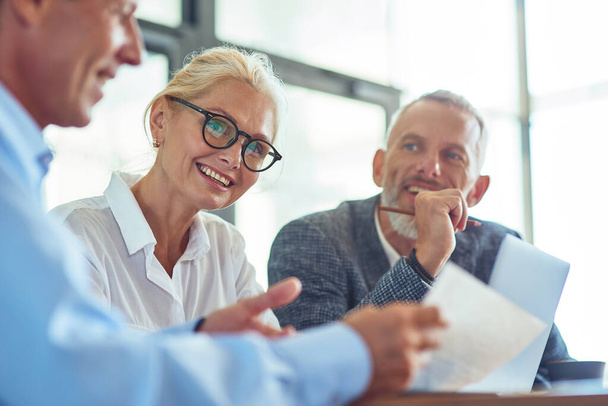 Mooie volwassen zakenvrouw dragen bril kijken naar de camera en glimlachen tijdens het bespreken van contract met partners in het moderne kantoor - Foto, afbeelding