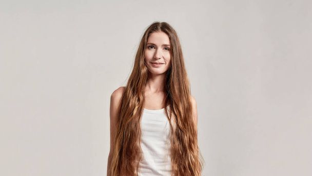 Portrait of young caucasian woman with long hair wearing white shirt smiling at camera while standing isolated over grey background - Foto, Bild