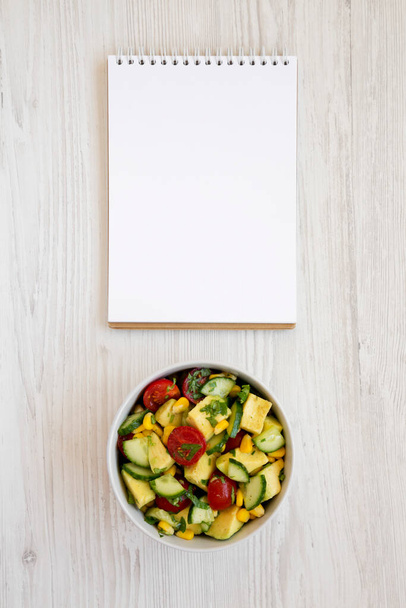 Fresh Avocado Tomato Salad in a bowl, blank notepad on a white wooden background, top view. Flat lay, overhead, from above.  - Photo, Image