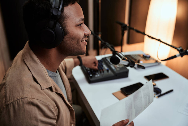 Retrato de um jovem apresentador de rádio masculino entrando ao vivo no ar, falando em microfone, segurando um papel de roteiro enquanto estava sentado em estúdio - Foto, Imagem