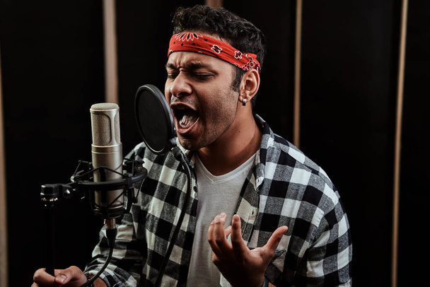 Portret van een jonge man, hiphop artiest die met gesloten ogen zingt in een condensator microfoon terwijl hij een lied opneemt in een professionele studio - Foto, afbeelding