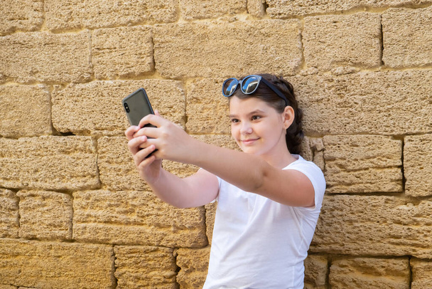 Stilvolles Teenager-Mädchen macht Selfie in der Stadt vor Steinmauer-Hintergrund. - Foto, Bild