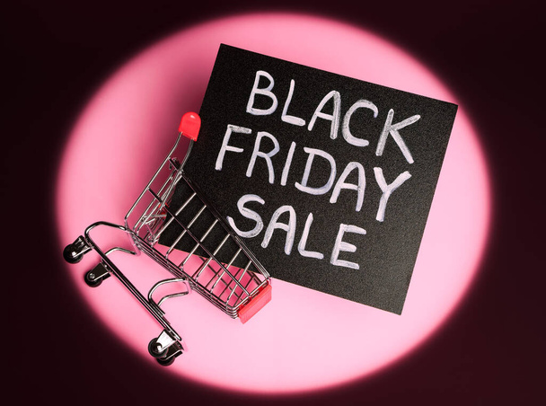 plano de pizarra de tiza negra con letras de tiza "venta de viernes negro" en un carrito de compras sobre un fondo rosa con viñeta circular oscura - Foto, imagen