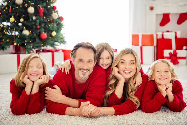Photo of full big family five people x-mas gathering three small kids hug hold hand cheeks lie floor carpet wear red jumper in living room evergreen tree lights giftboxes indoors - 写真・画像