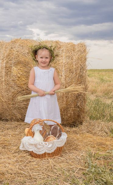 Το ξανθό κοριτσάκι με το στεφάνι στο κεφάλι κρατά ένα μπουκέτο σιτάρι στο χωράφι.. - Φωτογραφία, εικόνα