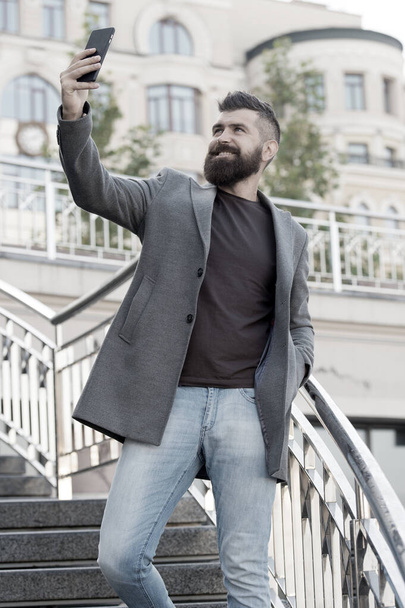 Hand selfie. Bearded man take selfie with smartphone outdoor. Businessman smile to selfie camera in mobile phone. Enjoying selfie session in business style. Modern life - 写真・画像