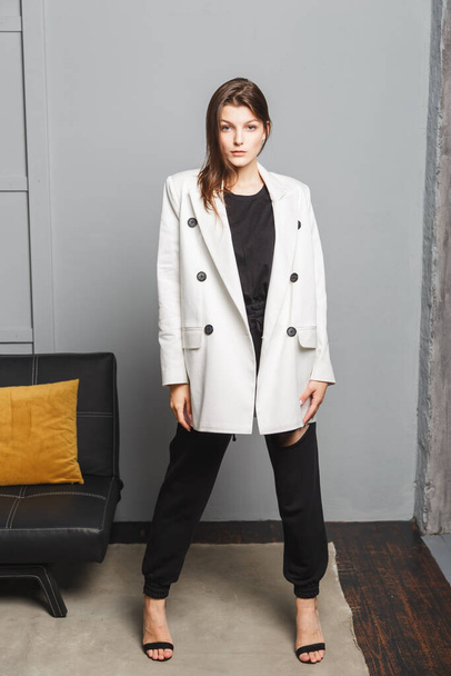 Young elegant woman in white oversize blazer and black ripped pants. Interior portrait - Photo, Image
