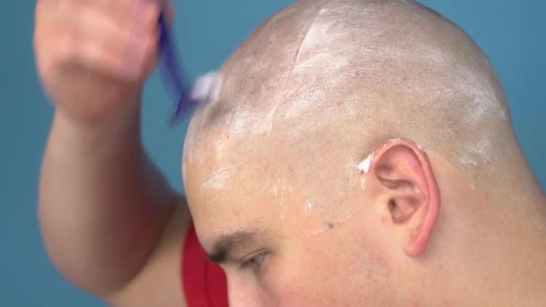 Holohlavý mladík si drze oholí hlavu. Muž si stříhá vlasy břitvou na modrém pozadí. - Záběry, video