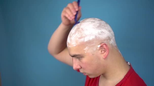 De kale jongeman scheert brutaal zijn hoofd. Een man knipt zijn haar met een scheermes op een blauwe achtergrond. - Video