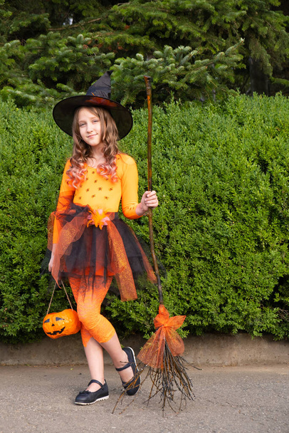 beautiful cute schoolgirl in witch halloween orange costume hold the broom and pumpkin. - Photo, Image