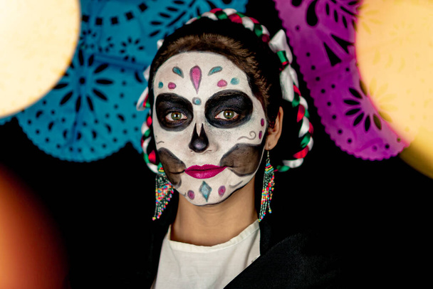 Μεξικανή γυναίκα με παραδοσιακή Ημέρα των Νεκρών μακιγιάζ, πίσω διακοσμημένα με πολύχρωμες λωρίδες από παραδοσιακό μεξικάνικο χαρτί και μαύρο φόντο. - Φωτογραφία, εικόνα